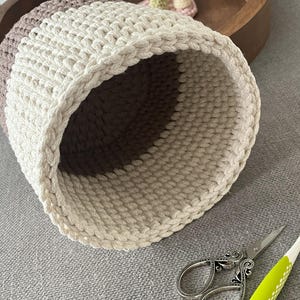 May include: A close-up of a crocheted basket with a brown and white colour scheme. The basket is made of thick, chunky yarn and has a round shape. The basket is sitting on a grey surface.