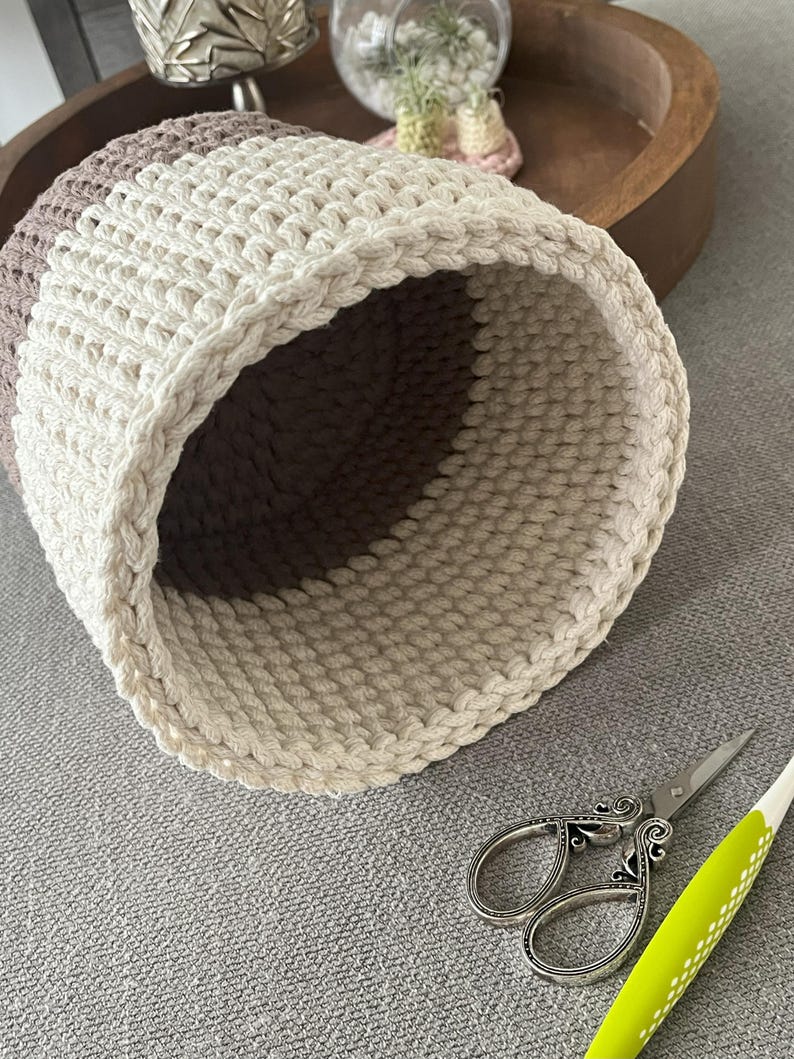 May include: A close-up of a crocheted basket with a brown and white colour scheme. The basket is made of thick, chunky yarn and has a round shape. The basket is sitting on a grey surface.