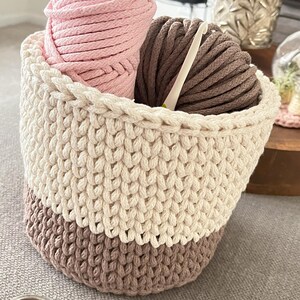 May include: A crocheted basket with a white top and brown bottom. The basket is filled with pink and brown yarn and a crochet hook.