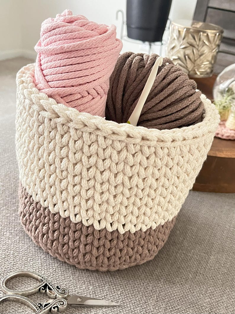May include: A crocheted basket with a white top and brown bottom. The basket is filled with pink and brown yarn and a crochet hook.