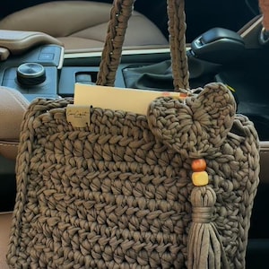 Może przedstawiać: Brązowa torba na ramię wykonana na szydełku z serduszkowym breloczkiem i frędzlem. Torba ma małą etykietę z napisem "Crochet Creations".