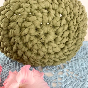 May include: A green crocheted round cushion with a textured surface. The cushion is made of fabric strips and is sitting on a blue crocheted surface.