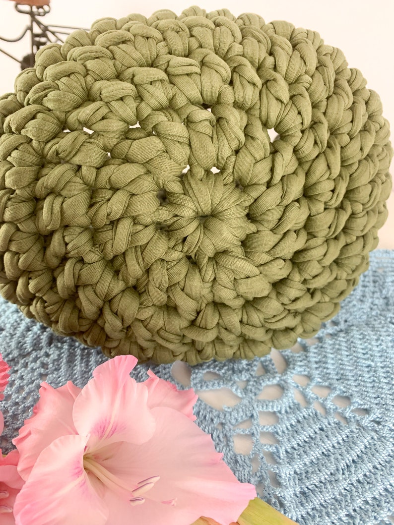 May include: A green crocheted round cushion with a textured surface. The cushion is made of fabric strips and is sitting on a blue crocheted surface.