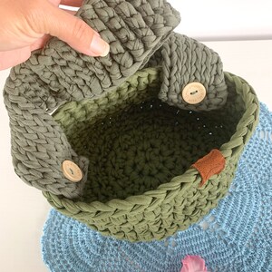 May include: A green crocheted basket with two fabric handles and a wooden button on each handle. The basket is sitting on a blue crocheted doily. The text on the button reads "Handmade with love".