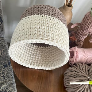 May include: A crocheted storage basket with a brown and white striped pattern. The basket is made of thick cotton rope and has a round shape.