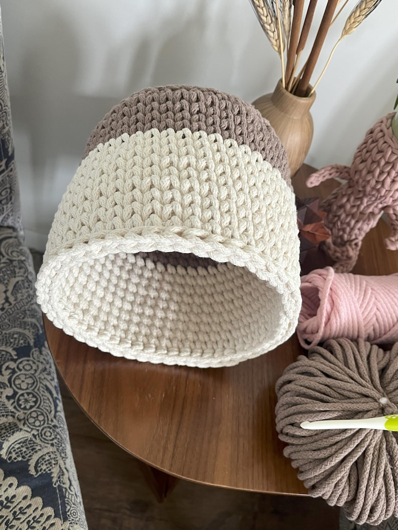 May include: A crocheted storage basket with a brown and white striped pattern. The basket is made of thick cotton rope and has a round shape.