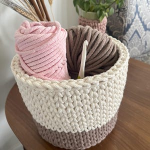 May include: A crocheted basket with a white and brown striped pattern, filled with pink and brown yarn and a crochet hook.
