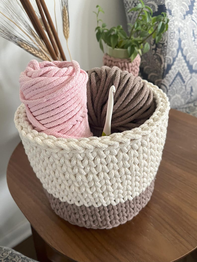 May include: A crocheted basket with a white and brown striped pattern, filled with pink and brown yarn and a crochet hook.