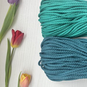 May include: Two spools of cotton rope, one teal and one dark teal, on a white surface.  There are three tulips in the background, one purple, one red, and one yellow.
