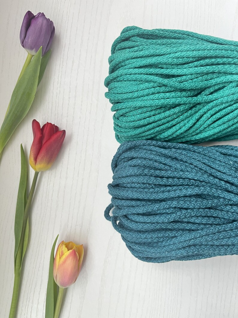 May include: Two spools of cotton rope, one teal and one dark teal, on a white surface.  There are three tulips in the background, one purple, one red, and one yellow.