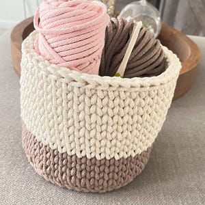 Peut inclure: Un panier en crochet à rayures blanches et brunes, rempli de fils roses et bruns et d'un crochet. Le panier est posé sur une surface brun clair.