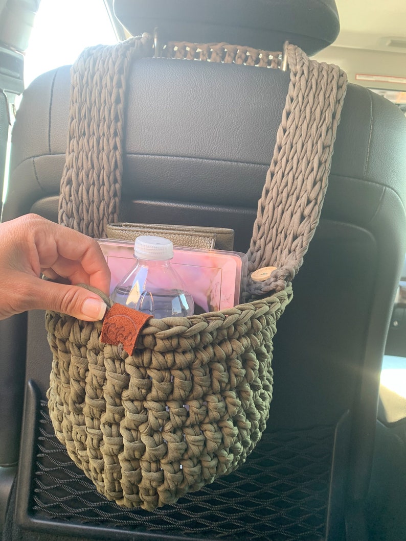 May include: A green crocheted hanging basket with a brown leather tag, attached to the headrest of a car seat. The basket contains a clear water bottle and a pink and white book.