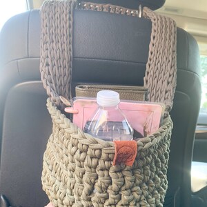 May include: A green crocheted hanging basket with a brown leather tag. The basket is filled with a water bottle, a pink notebook, and a brown leather wallet.