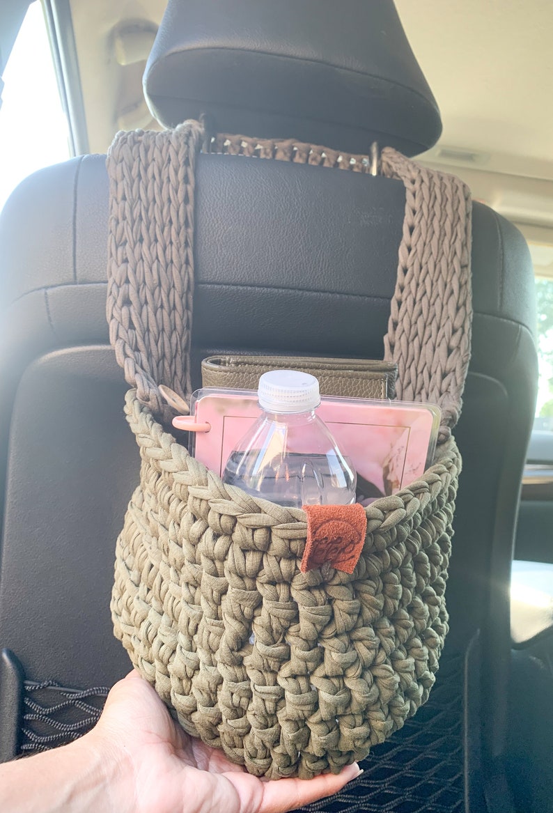 May include: A green crocheted hanging basket with a brown leather tag. The basket is filled with a water bottle, a pink notebook, and a brown leather wallet.
