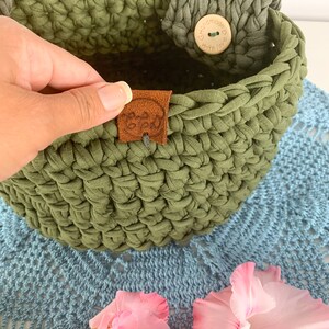 May include: A green crocheted basket with a brown leather tag that says "Handmade with love". The basket has a wooden button with the same text on it. The basket is sitting on a blue crocheted doily with pink flowers.