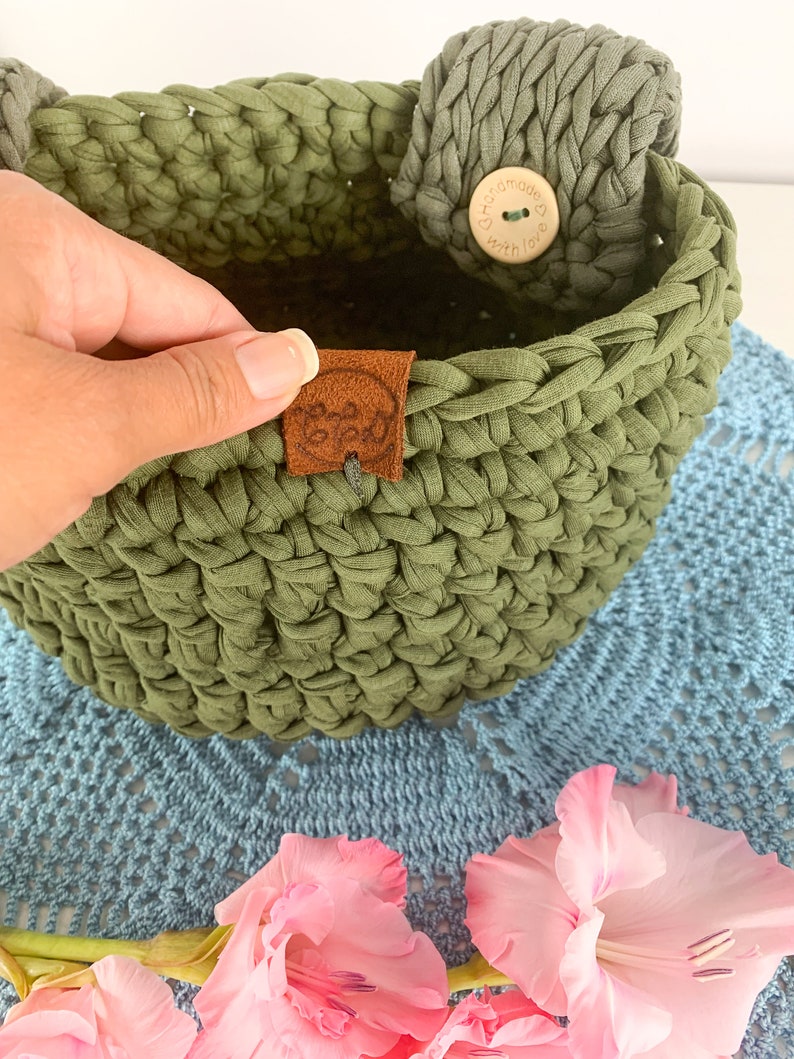 May include: A green crocheted basket with a brown leather tag that says "Handmade with love". The basket has a wooden button with the same text on it. The basket is sitting on a blue crocheted doily with pink flowers.