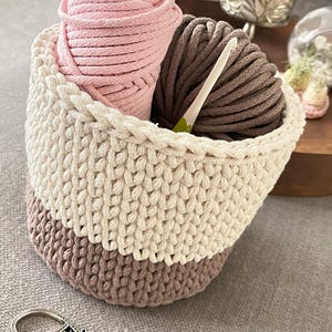 May include: A crocheted basket with a white, beige, and brown colour scheme. The basket is filled with pink and brown yarn balls and a crochet hook.
