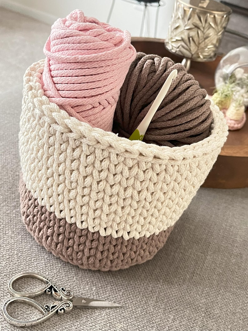 May include: A crocheted basket with a white, beige, and brown colour scheme. The basket is filled with pink and brown yarn balls and a crochet hook.