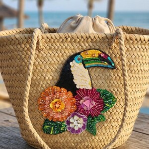 Puede incluir: Bolso tote de paja tejida con un tucán colorido y adornos florales. El tucán tiene un cuerpo negro, pecho blanco y un pico multicolor. El bolso tiene asas de cuerda y un cierre de cordón.
