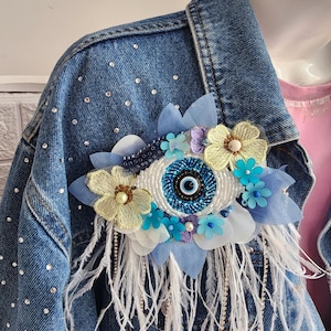 May include: A blue denim jacket with white feathers and a blue evil eye charm with white and yellow flowers.