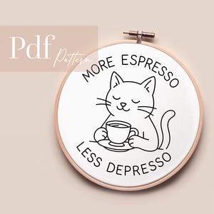 Könnte beinhalten: Ein weißer Stickrahmen mit einer Katzenillustration, die eine Kaffeetasse hält. Der Text lautet "MORE ESPRESSO LESS DEPRESSO" in schwarzer Schrift. Der Rahmen befindet sich vor einem neutralen Hintergrund.