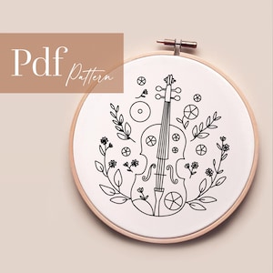 Puede incluir: Patrón de bordado de línea en blanco y negro de un violín con acentos florales. El patrón se muestra en una tela blanca en un aro de bordado redondo.