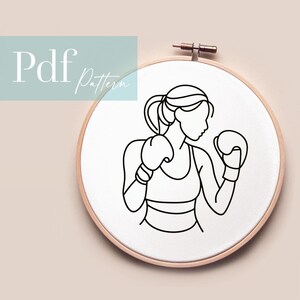 Könnte beinhalten: Eine schwarz-weiße Strichzeichnung einer Frau in Boxhaltung mit Boxhandschuhen ist auf einem weißen Stoff in einem hölzernen Stickrahmen zentriert. Der Text "Pdf Pattern" befindet sich in der oberen linken Ecke.
