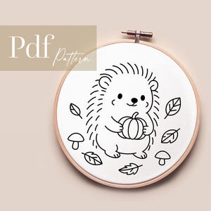 Könnte beinhalten: Ein Stickrahmen mit einer schwarz-weißen Strichzeichnung eines Igels, der einen Kürbis hält, umgeben von Blättern und Pilzen. Der Text "Pdf Pattern" befindet sich in der oberen linken Ecke.