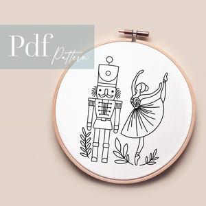 May include: Black and white line art embroidery pattern of a nutcracker and a ballerina. The pattern is in a circular hoop and includes a few sprigs of greenery.