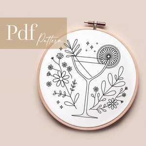 Könnte beinhalten: Schwarz-weißes Linienkunst-Stickmuster eines Cocktailglases mit einer Orangenscheibe und Blumen. Das Muster ist auf weißem Hintergrund und befindet sich in einem hölzernen Stickrahmen.