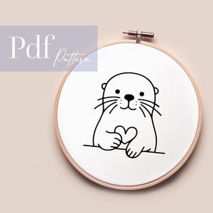 Könnte beinhalten: Schwarz-weiß-Zeichnung einer Otter, die ein Herz in ihren Pfoten hält. Die Otter lächelt und hat einen süßen Ausdruck. Die Zeichnung befindet sich auf einem weißen Hintergrund in einem hölzernen Stickrahmen.