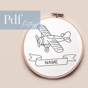 Könnte beinhalten: Eine Schwarzweiß-Zeichnung eines kleinen Flugzeugs mit einem Banner, auf dem "NAME" steht. Die Zeichnung befindet sich auf einem weißen Hintergrund in einem hölzernen Stickrahmen.