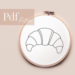 Könnte beinhalten: Ein Stickrahmen mit weißem Stoff und einer schwarzen Umrisszeichnung eines Croissants. Der Text "Pdf Pattern" ist sichtbar. Der Rahmen befindet sich vor einem hellrosa Hintergrund.