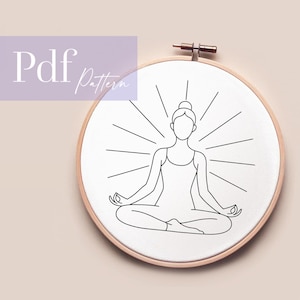 May include: A line drawing of a person in a yoga pose, radiating lines, is centered on a white fabric within a wooden embroidery hoop. The text "Pdf Pattern" is in the upper left corner. The design is simple and elegant.
