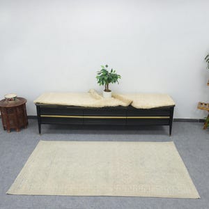 Puede incluir: Una alfombra rectangular de color claro con un patrón sutil está colocada sobre un suelo gris. Un mueble negro con ribetes dorados y una piel de oveja color crema están detrás de la alfombra. Una pequeña mesa de madera y un estante para plantas están a cada lado.