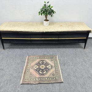 May include: A black mid-century modern style cabinet with three drawers and gold accents. The cabinet has a cream-colored, shaggy top. A small, square, patterned rug in pink, gray, and cream is on the floor. A small potted plant sits on top of the cabinet.