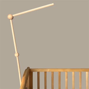 May include: A light-colored wooden crib with a matching mobile arm. The mobile arm is made of wood and has a ball joint and a horizontal bar at the top. The crib has vertical slats and a curved headboard.