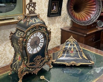 Könnte beinhalten: Eine antike schwarz-goldene Uhr mit floralen Akzenten und einem weißen Zifferblatt. Ein passendes Dekorationsteil befindet sich daneben. Ein Schlüssel liegt auf der grünen Oberfläche. Die Szene umfasst einen Vintage-Fernseher und ein Grammophon.