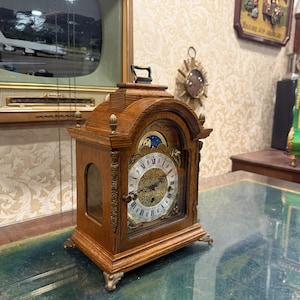 Puede incluir: Un reloj antiguo de madera con una esfera detallada, números romanos y manecillas ornamentadas. El reloj tiene un acabado marrón oscuro, un asa en la parte superior y detalles metálicos decorativos. El reloj está sobre una superficie de vidrio.