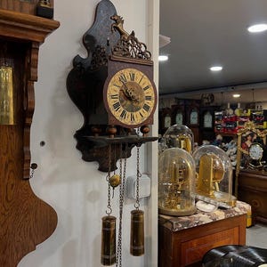Pode incluir: Um relógio de parede antigo em madeira escura, com um mostrador redondo e algarismos romanos. O relógio tem entalhes decorativos e pesos suspensos. Está montado numa parede branca, com outros relógios e objetos antigos ao fundo.