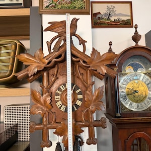 Könnte beinhalten: Eine Vintage-Kuckucksuhr aus Holz mit geschnitzten Blättern und einem Vogel, der obenauf sitzt. Das Zifferblatt der Uhr hat römische Ziffern und dekorative Zeiger. Die Uhr ist etwa 76 cm hoch.