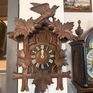 May include: A dark brown wooden cuckoo clock with a carved bird perched on top. The clock face has Roman numerals and decorative leaf accents. The clock is approximately 12 inches wide.