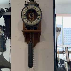 May include: A vintage wall clock with a wooden frame and a round face. The clock face has Roman numerals and decorative floral accents. A pendulum and weights hang below. The clock is mounted on a white wall.