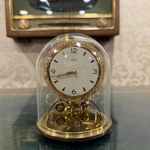 May include: A vintage gold-toned clock with a white face, black numerals, and gold hands, encased in a clear glass dome. The clock features a decorative gold rope-like trim and a pendulum base. The clock is a classic decorative item.