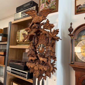 Könnte beinhalten: Eine detaillierte Kuckucksuhr aus Holz mit einem Adler, der obenauf sitzt. Die Uhr hat geschnitzte Blätter, Hirsche und ein kreisförmiges Zifferblatt mit römischen Ziffern. Die Uhr ist dunkelbraun und hat zwei hängende Gewichte.