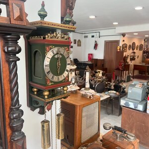 Könnte beinhalten: Eine antike Holzuhr mit grünem Zifferblatt und goldenen Akzenten. Die Uhr hängt an einer Wand in einem Geschäft mit anderen antiken Gegenständen.