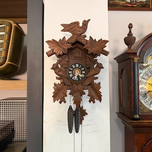 May include: A wooden cuckoo clock with a carved bird on top and two birds on the sides of the clock face. The clock face has Roman numerals and decorative leaves. Two pinecone-shaped weights hang below.