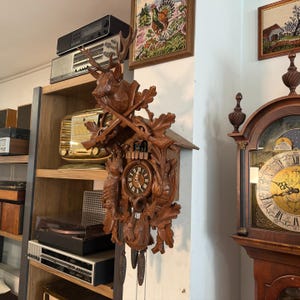 Könnte beinhalten: Eine detaillierte Kuckucksuhr aus Holz mit geschnitzten Hirschgeweihen, Blättern und einem Vogel. Das Zifferblatt der Uhr hat römische Ziffern. Die Uhr ist an einer weißen Wand montiert, mit anderen Vintage-Gegenständen im Hintergrund.