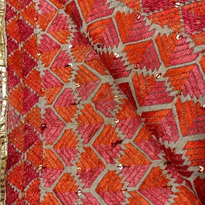 Könnte beinhalten: Nahaufnahme eines Textils mit aufwendiger Stickerei in Orange-, Rosa- und Rottönen. Das Design weist geometrische Muster und kleine, glänzende Verzierungen auf. Ein goldener Rand säumt den Rand.