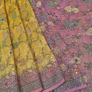 May include: A textile fabric featuring a gradient from yellow to pink, decorated with an embroidered floral pattern in dark green and gold threads. The design is accented with small silver sequins and scattered round purple and yellow rhinestones.